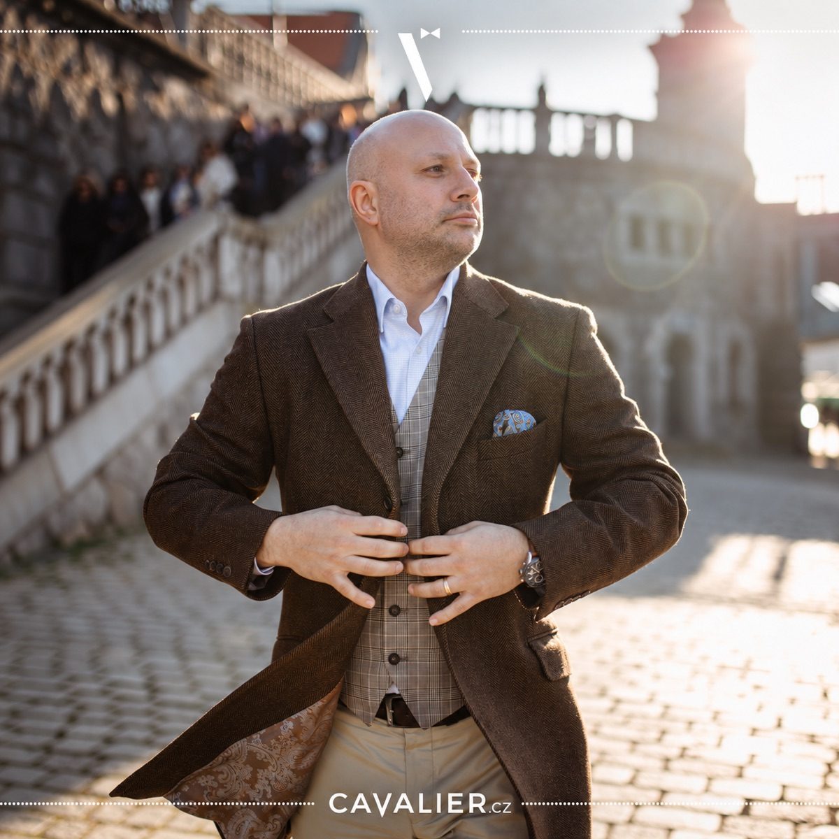 Man in a long brown coat over a checked waistcoat and white shirt.