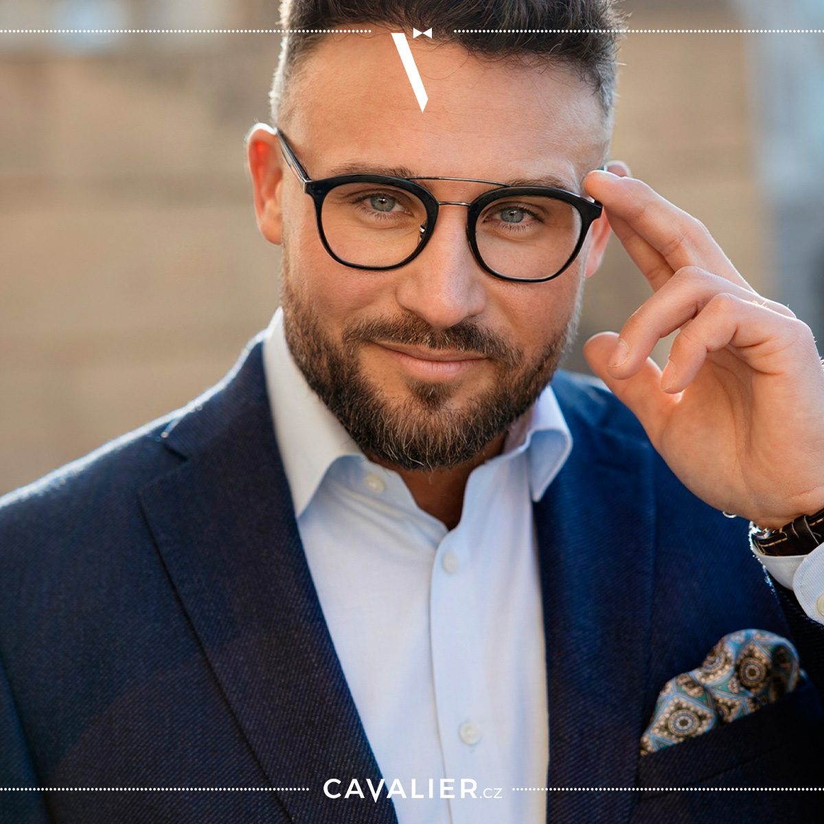 Portrait of a man with glasses and a groomed beard in a navy Cavalier made-to-measure jacket and light shirt