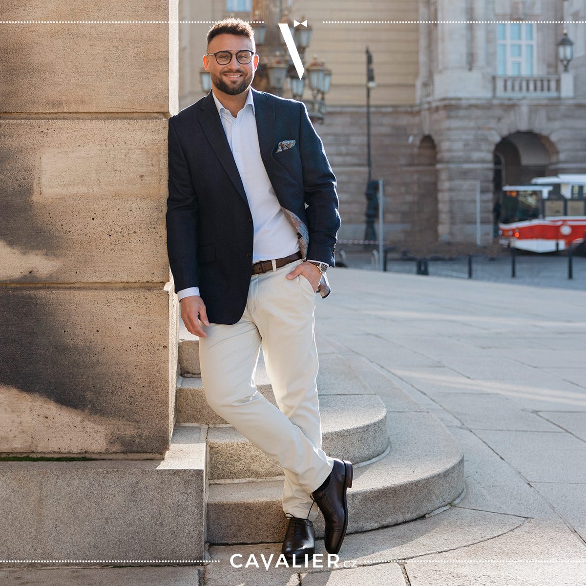 A man leaning against a city column in a navy Cavalier separate jacket, patterned pocket square, open-collar white shirt and light chinos with a brown belt