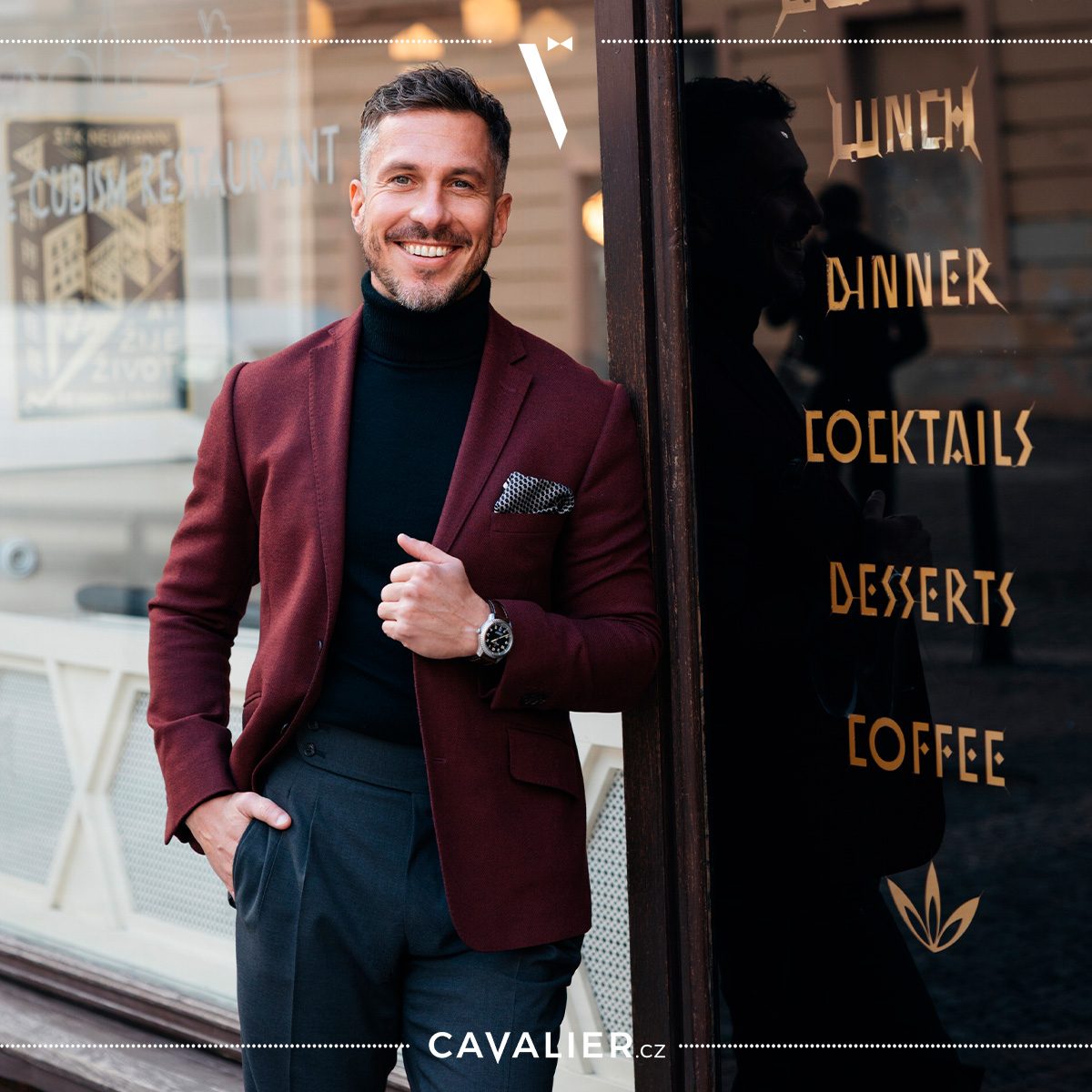 Man in a burgundy jacket and black roll-neck beside a glass entrance.