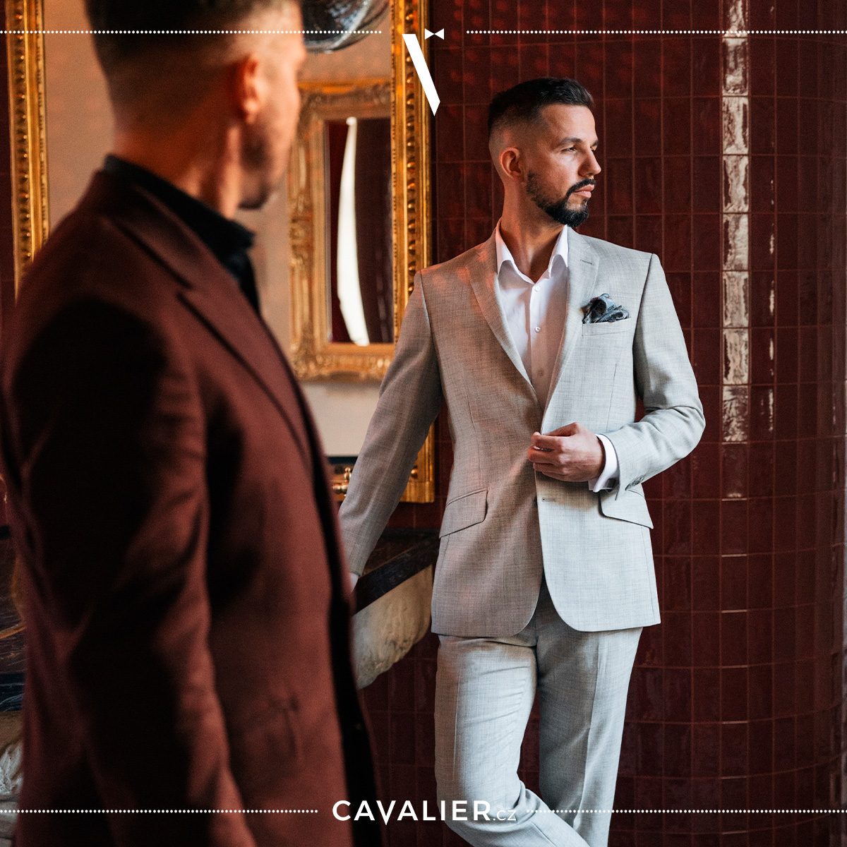 Two men in an interior: a burgundy jacket in the foreground and a light-grey suit behind.