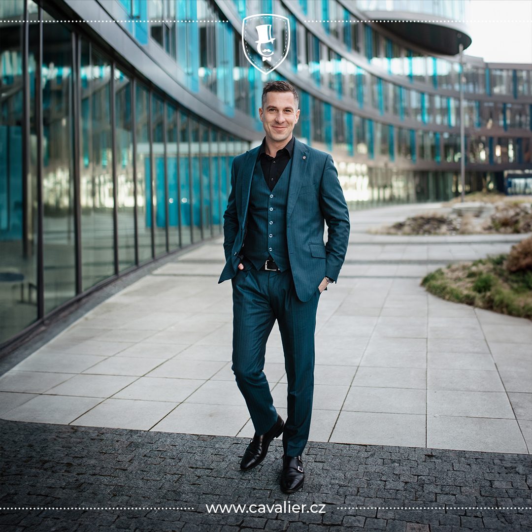 Man in a dark teal suit and waistcoat standing outside a glass-fronted building.