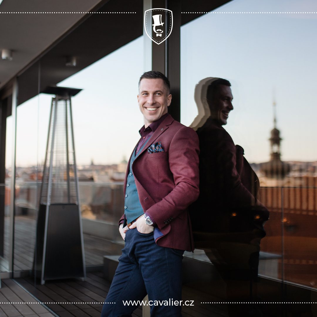 A smiling man in a burgundy Cavalier made-to-measure jacket, dark green waistcoat, dark shirt and jeans, combining colour and textured fabrics