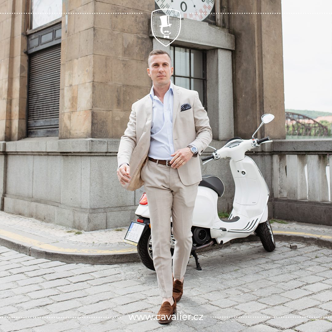 Man in a beige suit and brown shoes walking beside a white scooter.