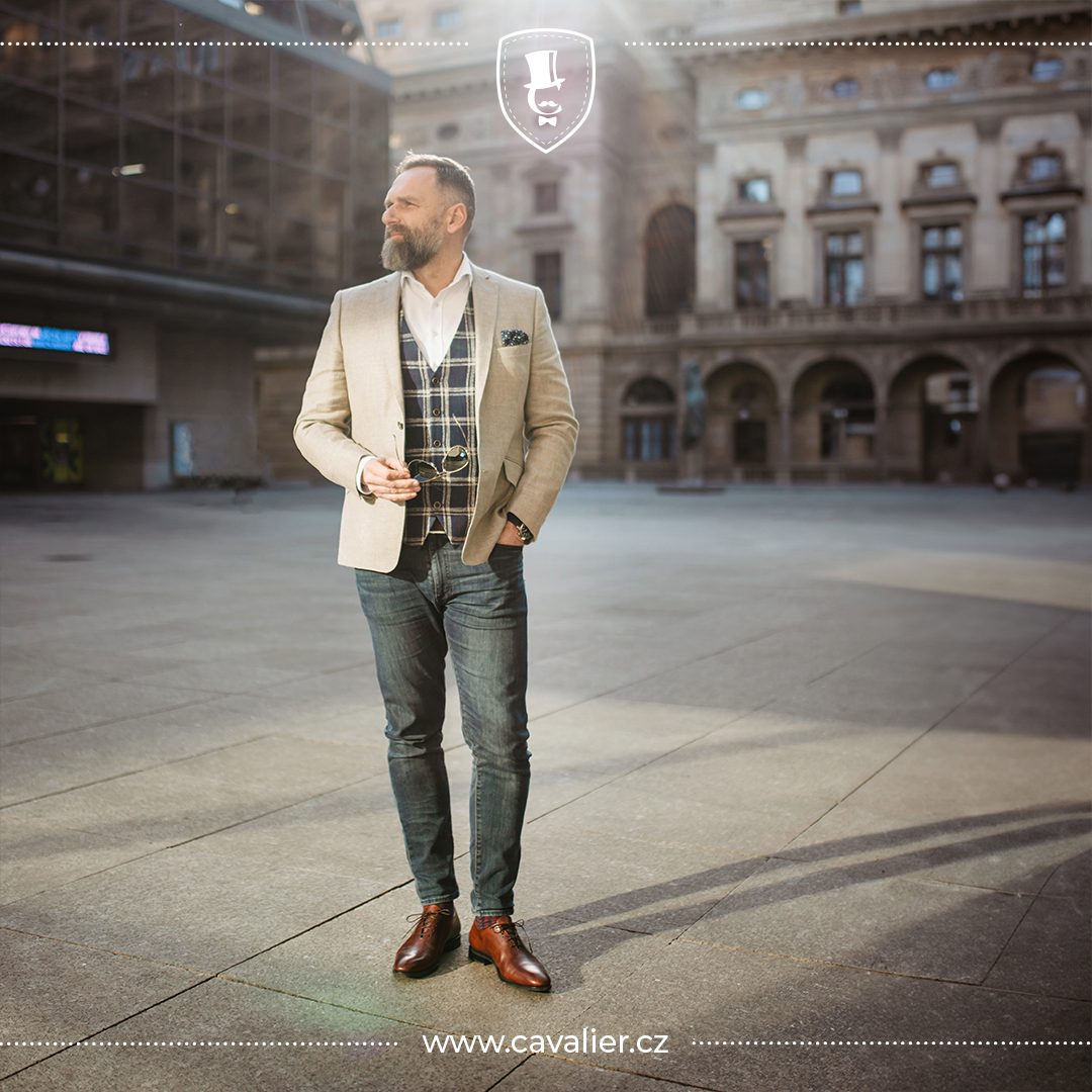 A man in a smart casual outfit: a light Cavalier made-to-measure wool jacket, checked waistcoat, open-collar white shirt and dark jeans
